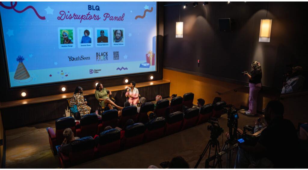 BLQ Disruptors Panel at a small theater, speakers seated, audience attentively watching, screen shows panelist names and sponsors