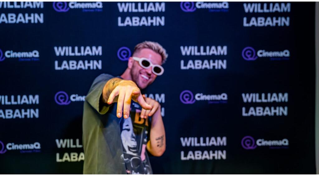 William Labahn pointing playfully, wearing sunglasses, against a backdrop reading "CinemaQ Denver Film" and "WILLIAM LABAHN"