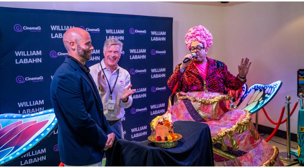 Three men at a CinemaQ event, one in colorful attire speaking into a microphone