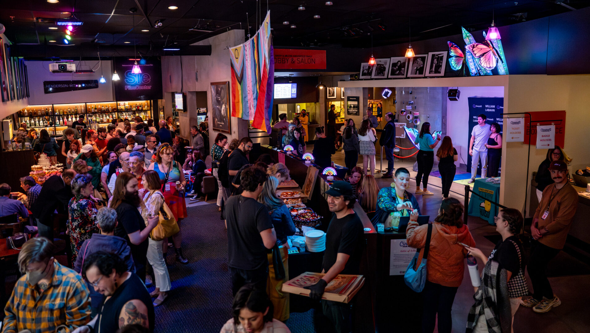 Busy cinema lobby with crowds, colorful flags, rainbow decorations, and a bar