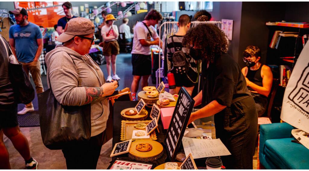 People shopping at a market booth, vendor organizing items, small crowd in the background