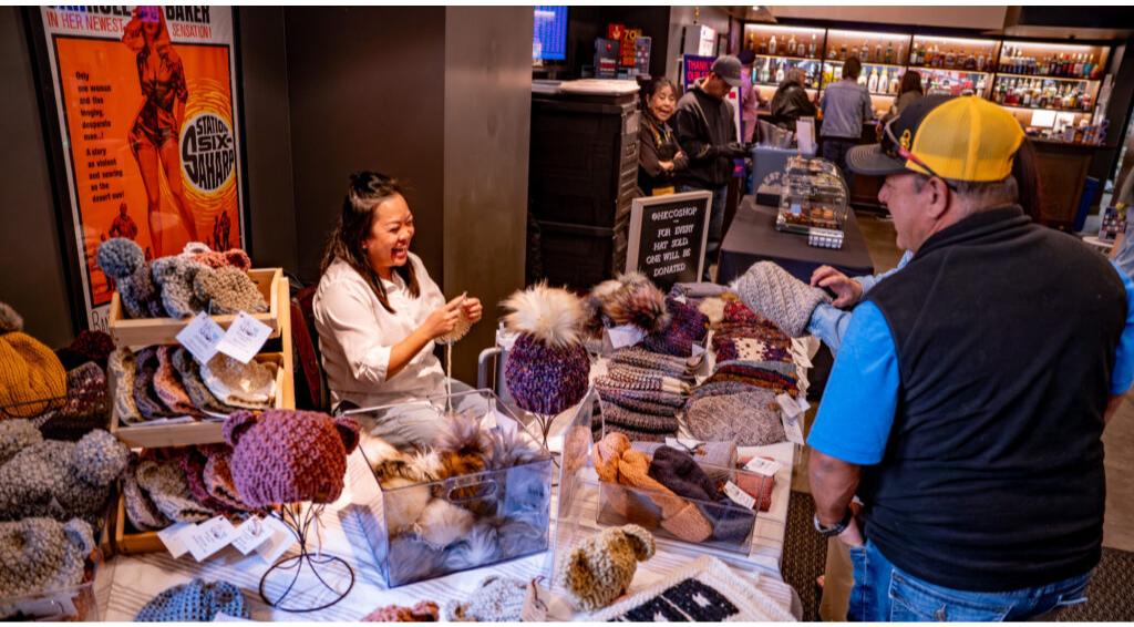 Vendor DBFF25_Day2_BNGU-31 interacting with customer, displaying knitted hats and scarves, sign reads "One for You, One Will Be Donated."