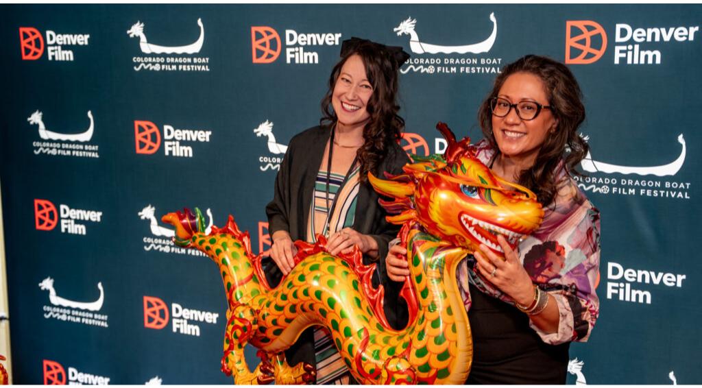 Two smiling women holding an inflatable dragon at Colorado Dragon Boat Film Festival, Denver Film logos in background