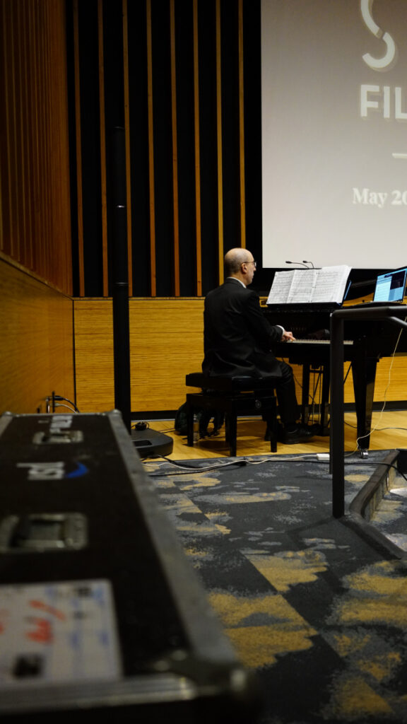 DSC00340.jpeg playing piano in dim auditorium; May 2019 on screen