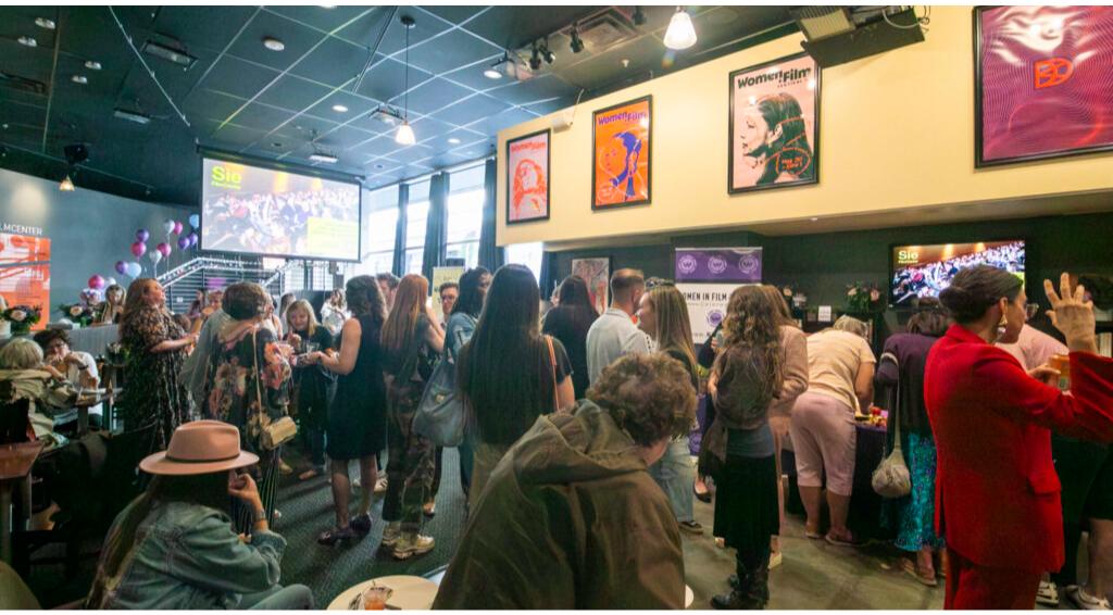Large indoor gathering with people socializing, surrounded by film posters, and a screen displaying "Sie."