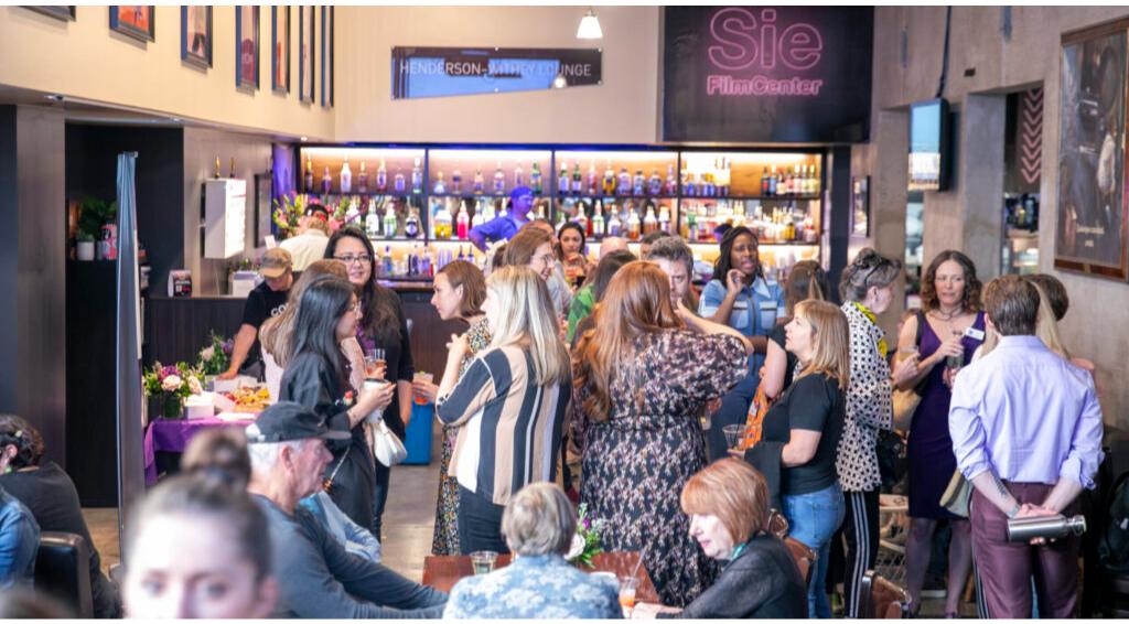 Crowded event at Sie FilmCenter with people socializing near a bar, various drinks, and floral decorations