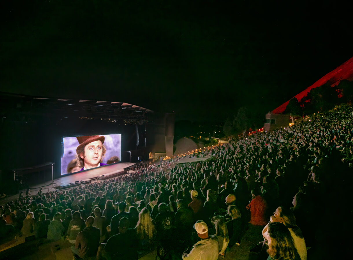 Outdoor amphitheater at night with a large screen showing a Willy Wonka scene, attended by a large crowd