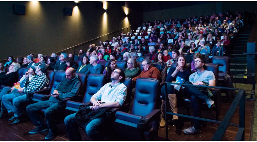 Audience seated in a theater, watching a performance or screening, diverse group, some eating snacks