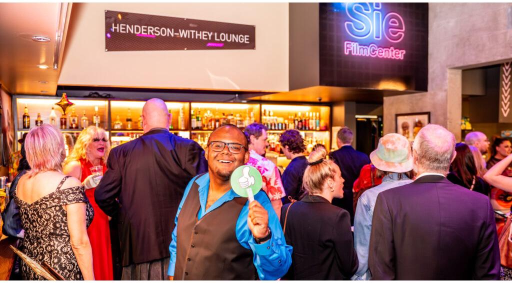 Man in blue shirt at crowded Henderson-Withey Lounge event, smiling and holding a green card