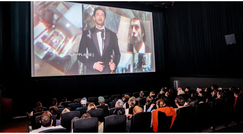 Audience watches Oscar speech on movie theater screen displaying man in tuxedo holding an award, captioned "[APPLAUSE]."