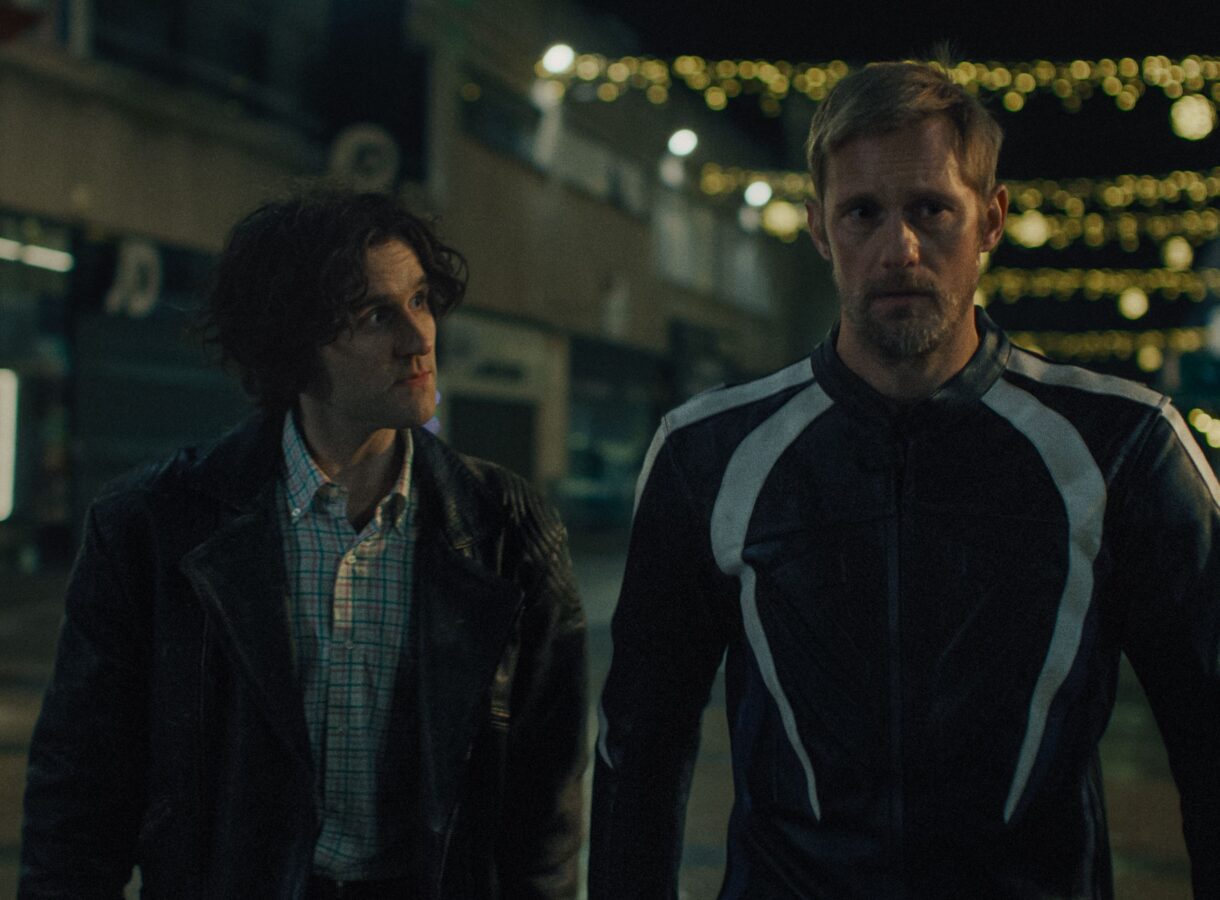 Two men walking at night, one in a leather jacket, the other in a motorcycle suit, under string lights