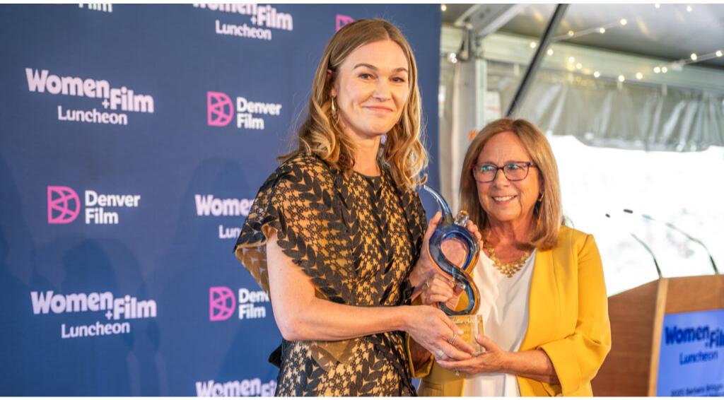 Julia Stiles holding an award at the Women+Film Luncheon with a smiling companion in a yellow blazer