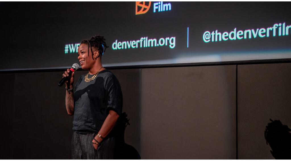 Woman speaking into a microphone at a film event, smiling. Background displays text: "Denver Film," "denverfilm.org," "@thedenverfilm"