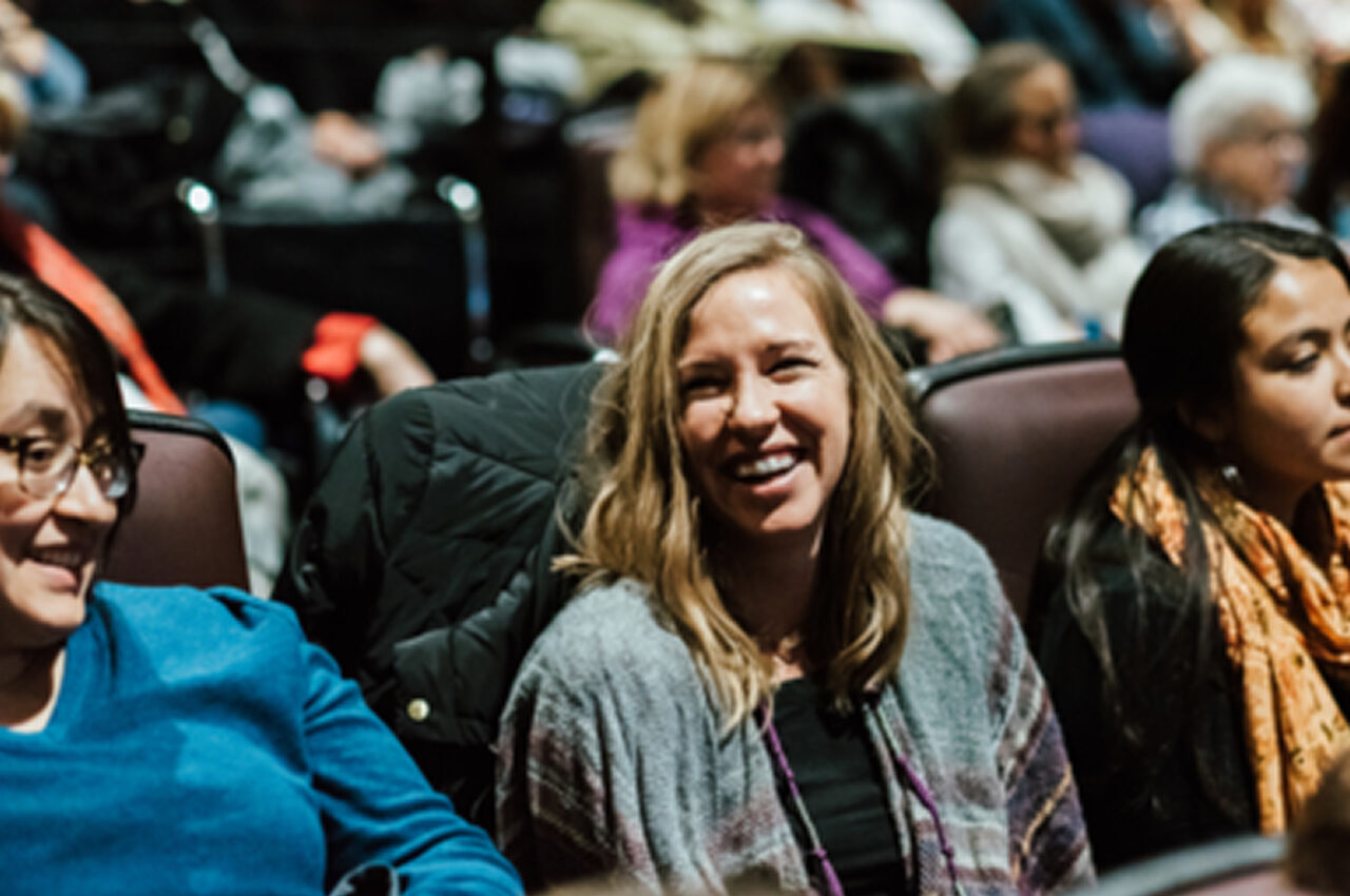 People seated in an audience, focusing on a smiling woman in conversation