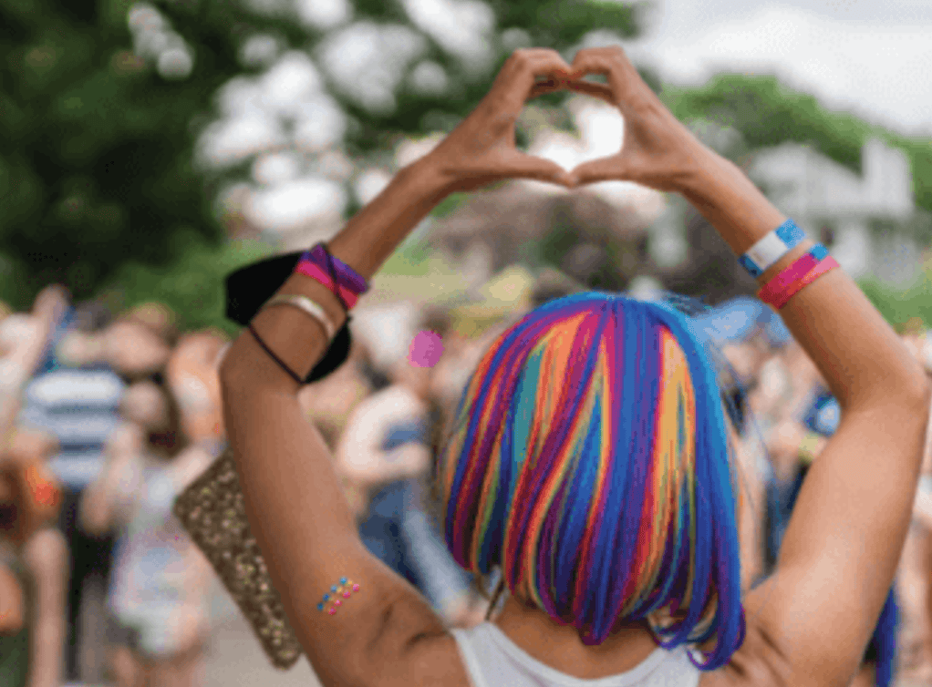 Person with rainbow wig forming heart with hands at a colorful outdoor event