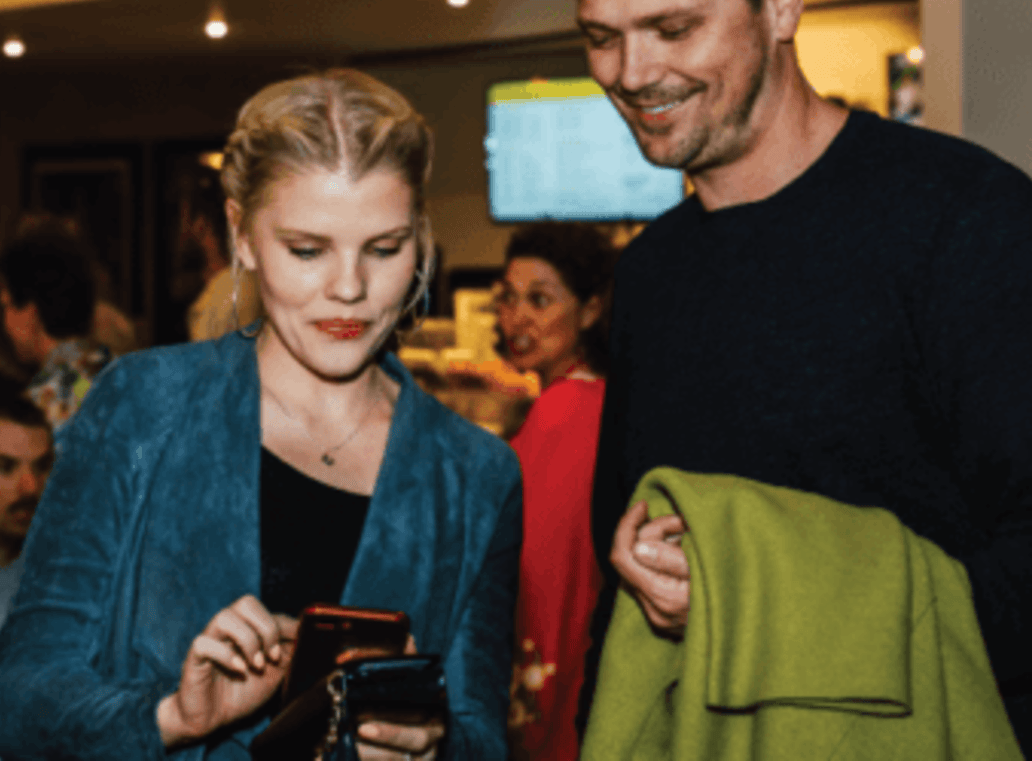 Woman in blue jacket using smartphone, man holds green jacket nearby, both smiling in a social setting