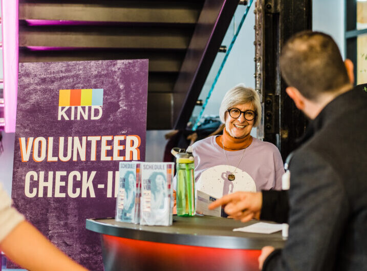 Volunteer check-in desk with "KIND" sign, smiling woman interacting with man, flower arrangement nearby