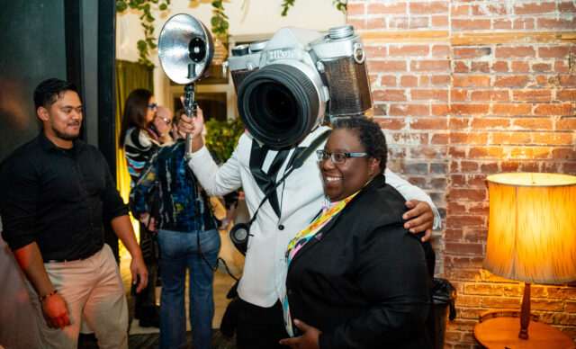A person in a camera helmet poses with a guest at a party, while people socialize in the background