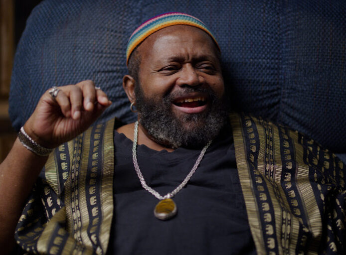 Carl Bean smiling warmly, wearing a patterned garment and colorful cap, seated in a cozy chair