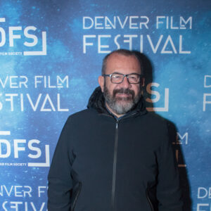 Man standing at Denver Film Festival, blue backdrop with "DFS" and "DENVER FILM FESTIVAL" text