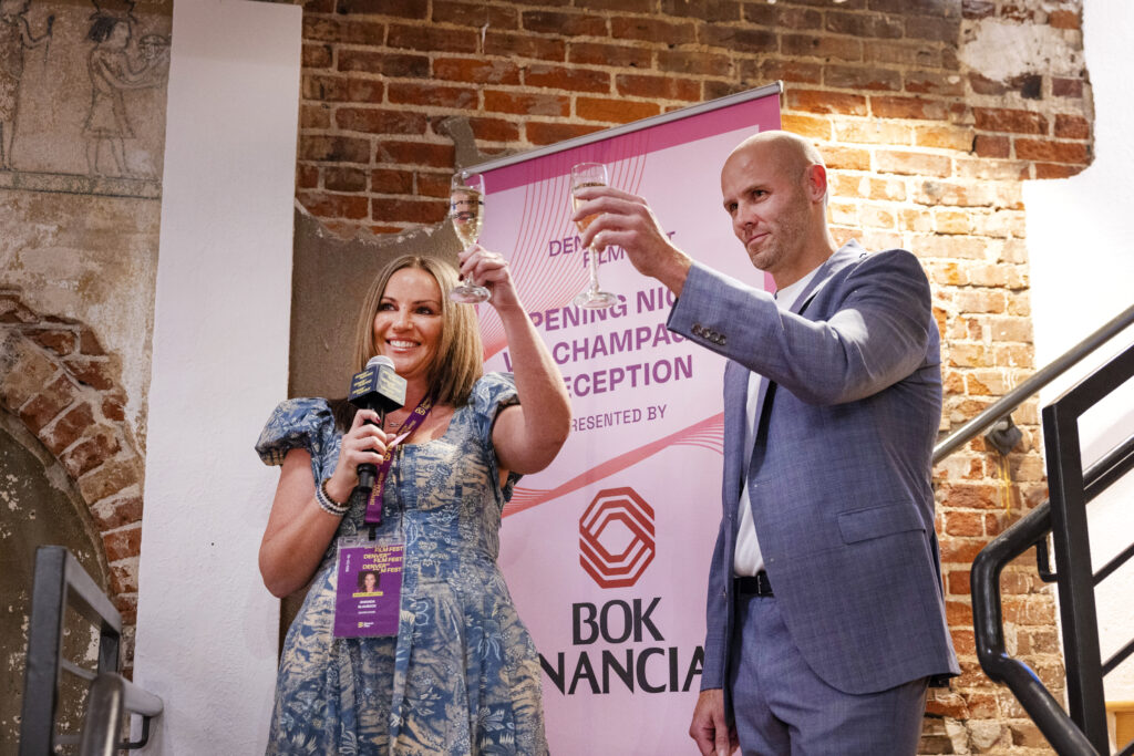 Rachel O'Driscoll smiling, holding a microphone and champagne, next to a man at an event with a "BOK Financial" banner