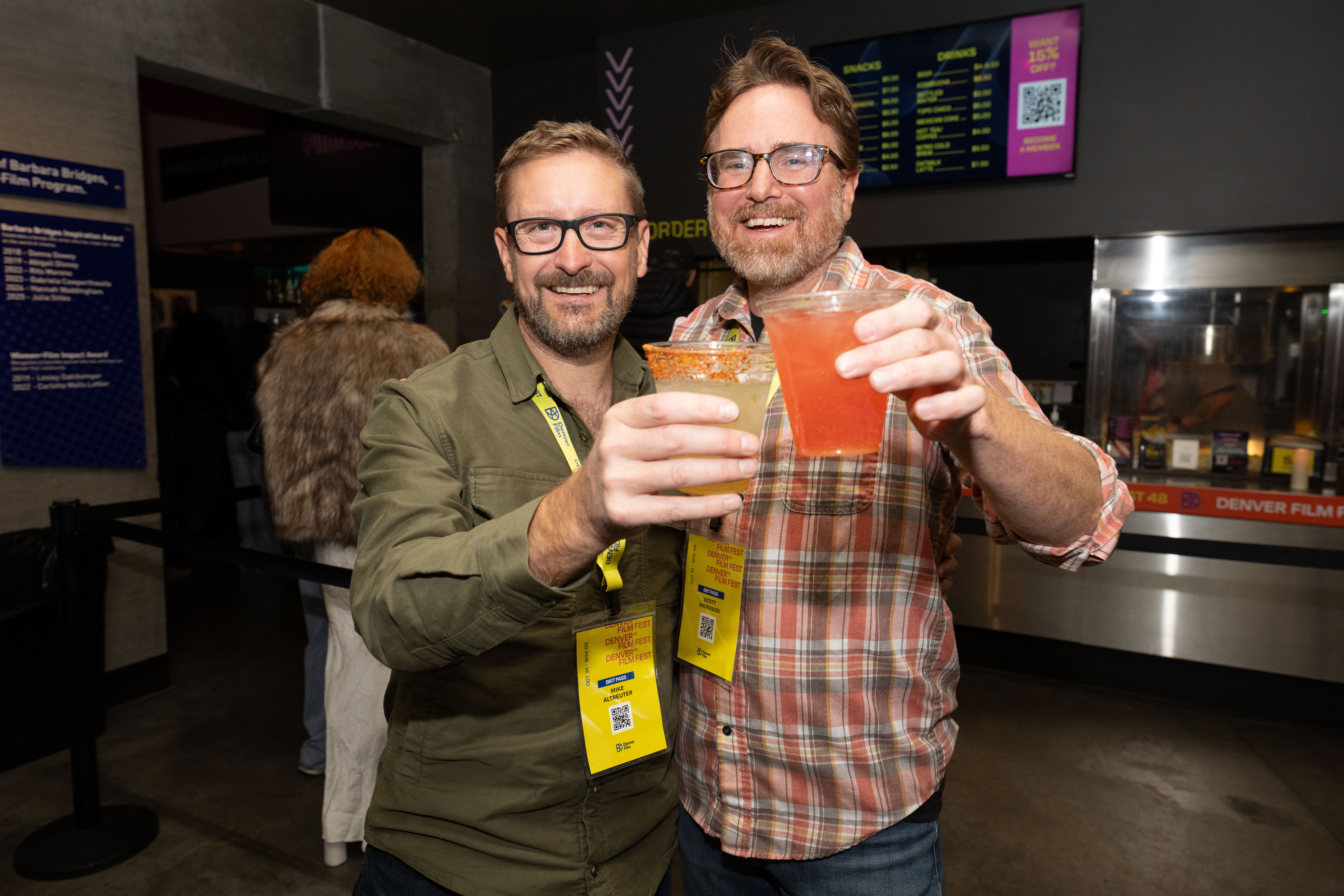 Two men smiling and toasting with drinks at an event, wearing badges