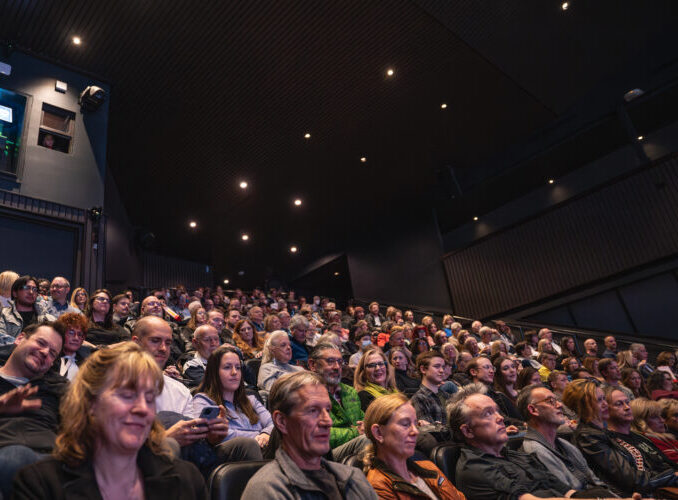 Audience seated in a large theater, focused on a presentation