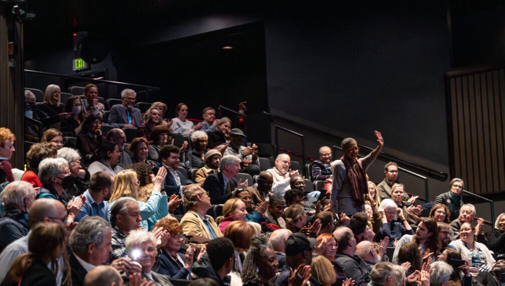 Audience clapping as a person stands and waves during an event