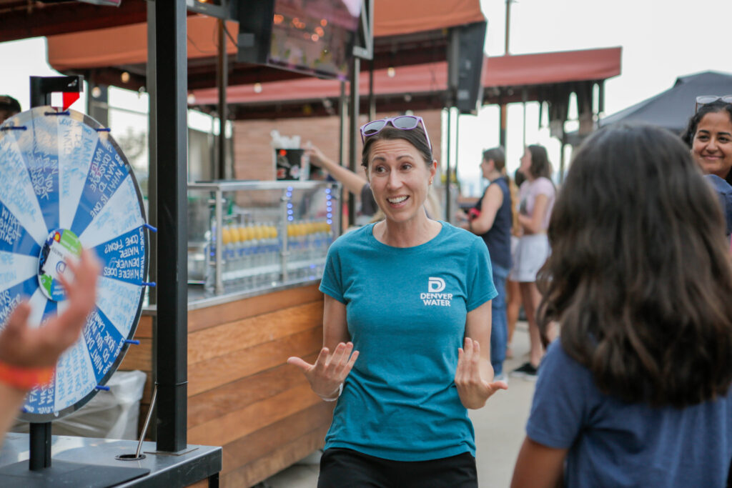 Person in a Denver Water shirt speaking animatedly near a prize wheel