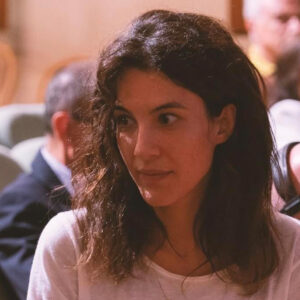 Carolina sitting attentively in a softly lit room with several people in the background