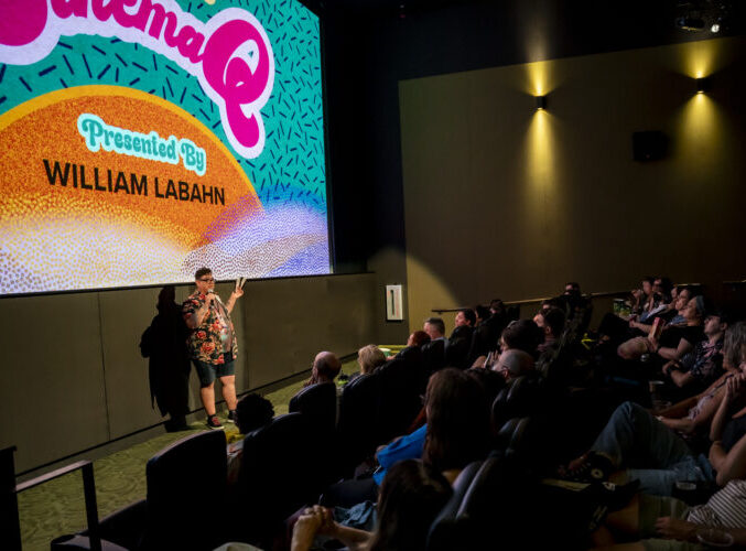 Person speaking to audience in theater; screen displays "CinemaQ Presented By William Labahn."