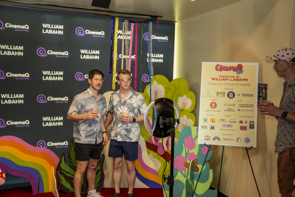 Two people at CinemaQ event, smiling with drinks, floral shirts; step-and-repeat backdrop, vibrant decorations, sponsor board visible