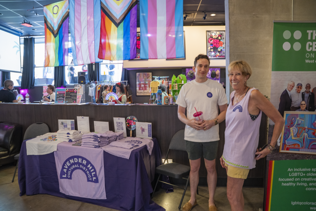 Two people at a Pride event booth, rainbow flags, Lavender Hill Cultural District table, promotional materials displayed
