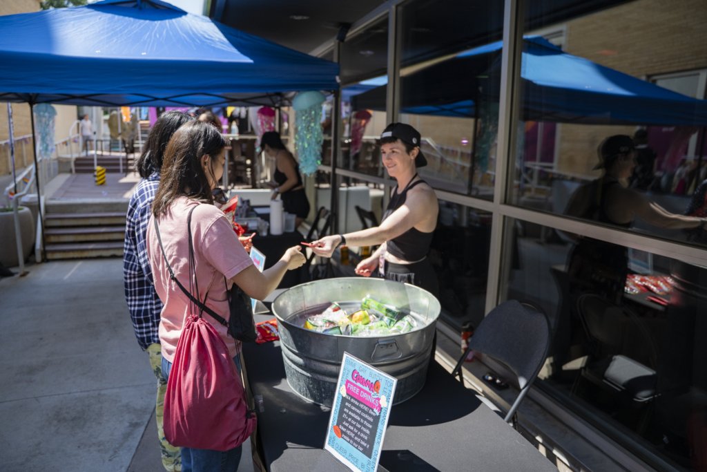 Person in tank top exchanging items with individuals at outdoor booth with blue tents