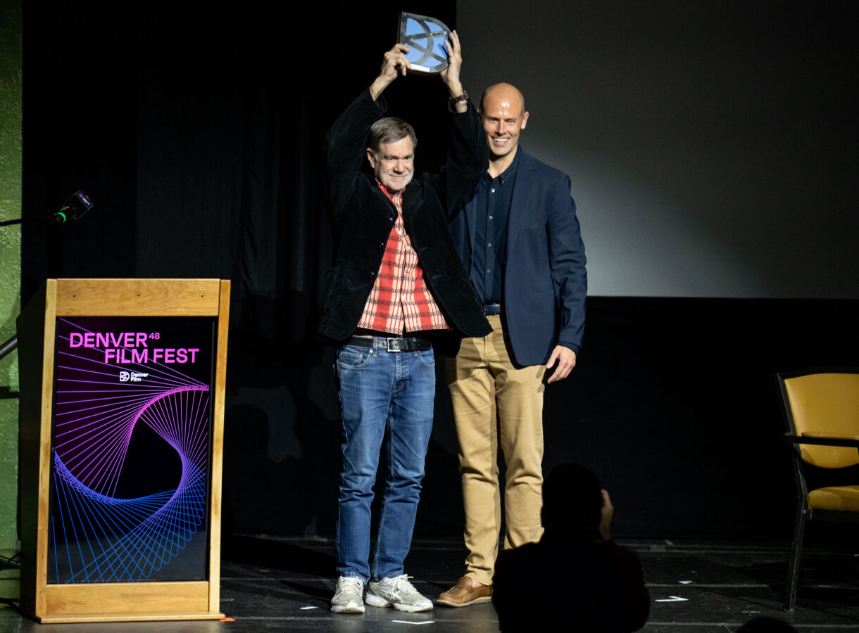 Two people on stage; one raises an award at Denver Film Fest