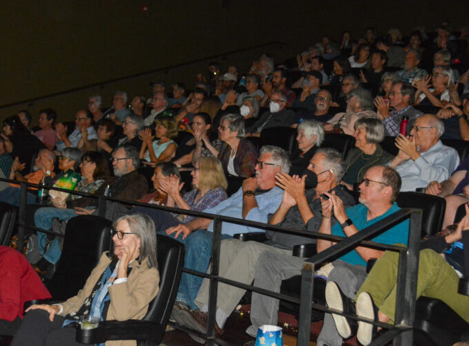Audience applauding in a dimly lit theater, showing diverse reactions and interest