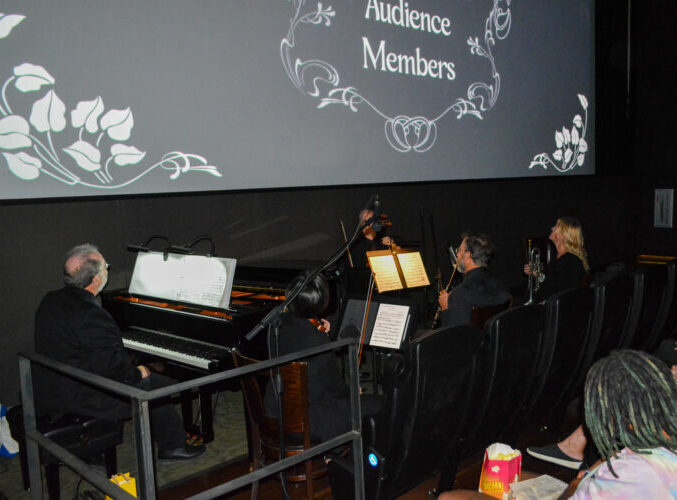 Musicians performing in a theater beside audience, screen displays "Audience Members" with floral design