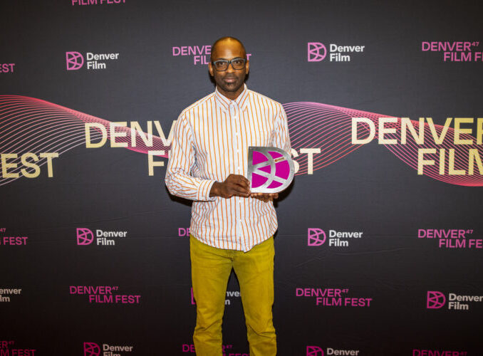 DeWitt holding award at Denver Film Fest backdrop, wearing striped shirt and yellow pants