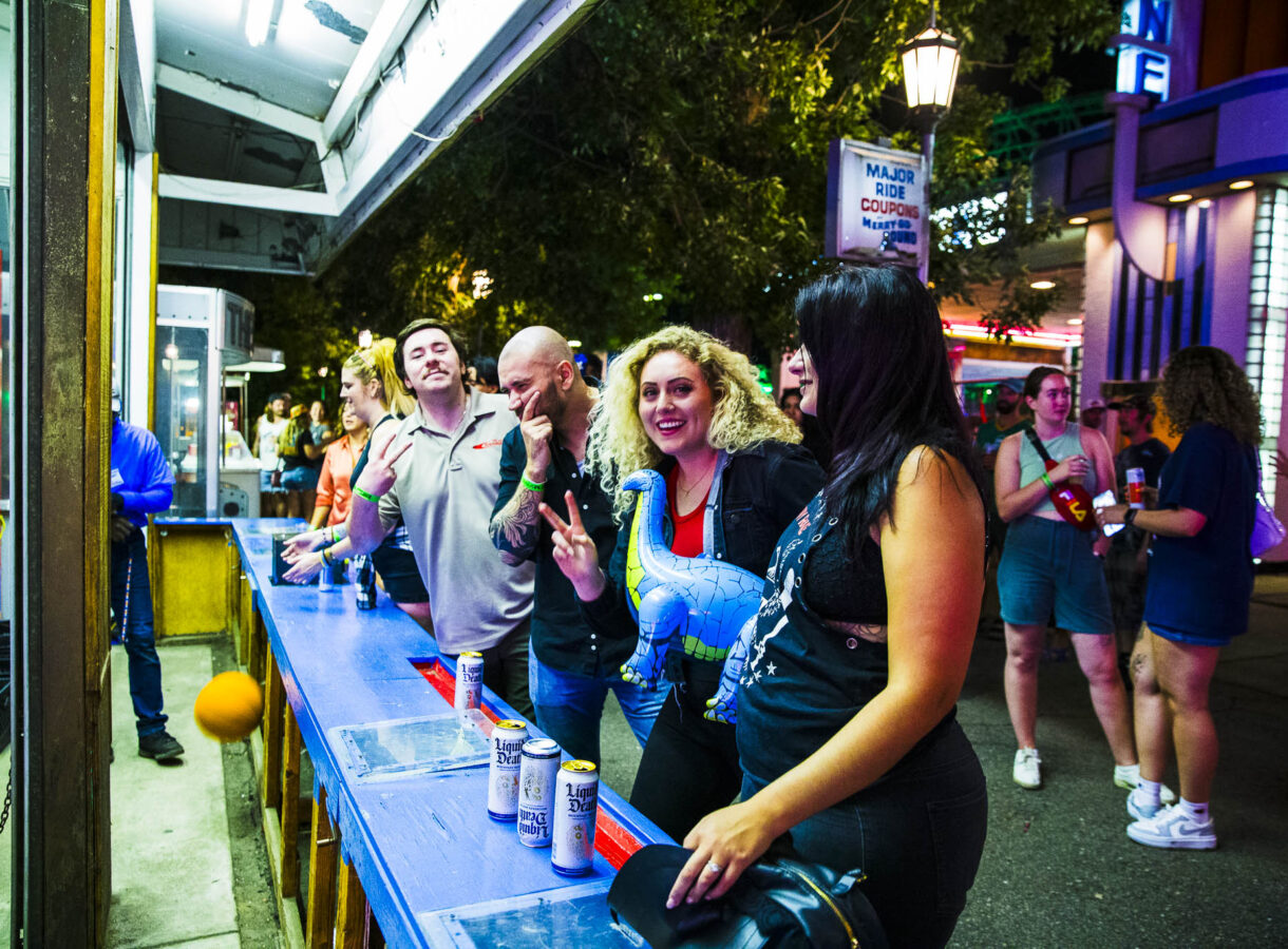 People at a lively outdoor event, person with curly hair smiling, holding inflatable dinosaur and making a peace sign