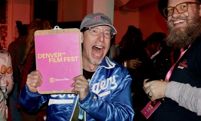 Man in Dodgers jacket excitedly holding "Denver 48 Film Fest" clipboard, smiling bearded man beside him