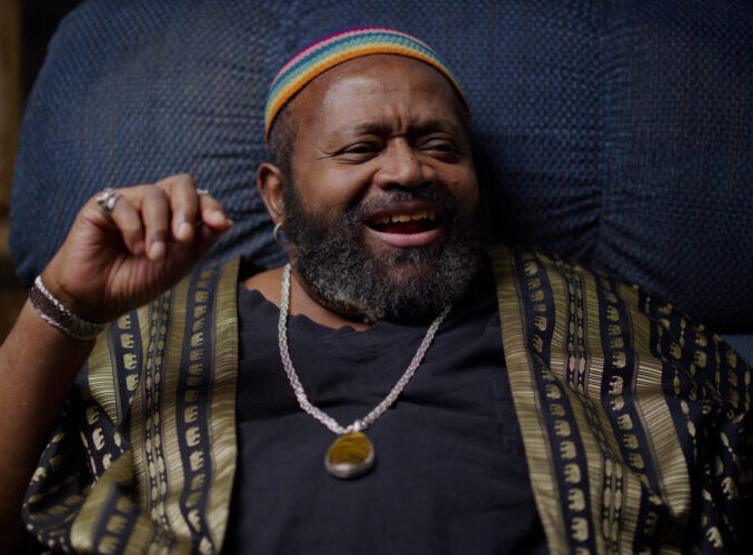 An individual from IWBTW, wearing a colorful cap and patterned robe, smiling while seated