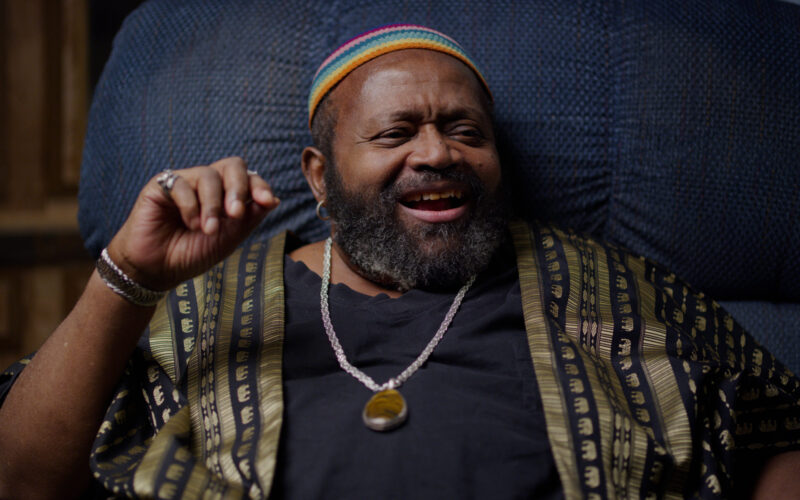 An individual from IWBTW, wearing a colorful cap and patterned robe, smiling while seated