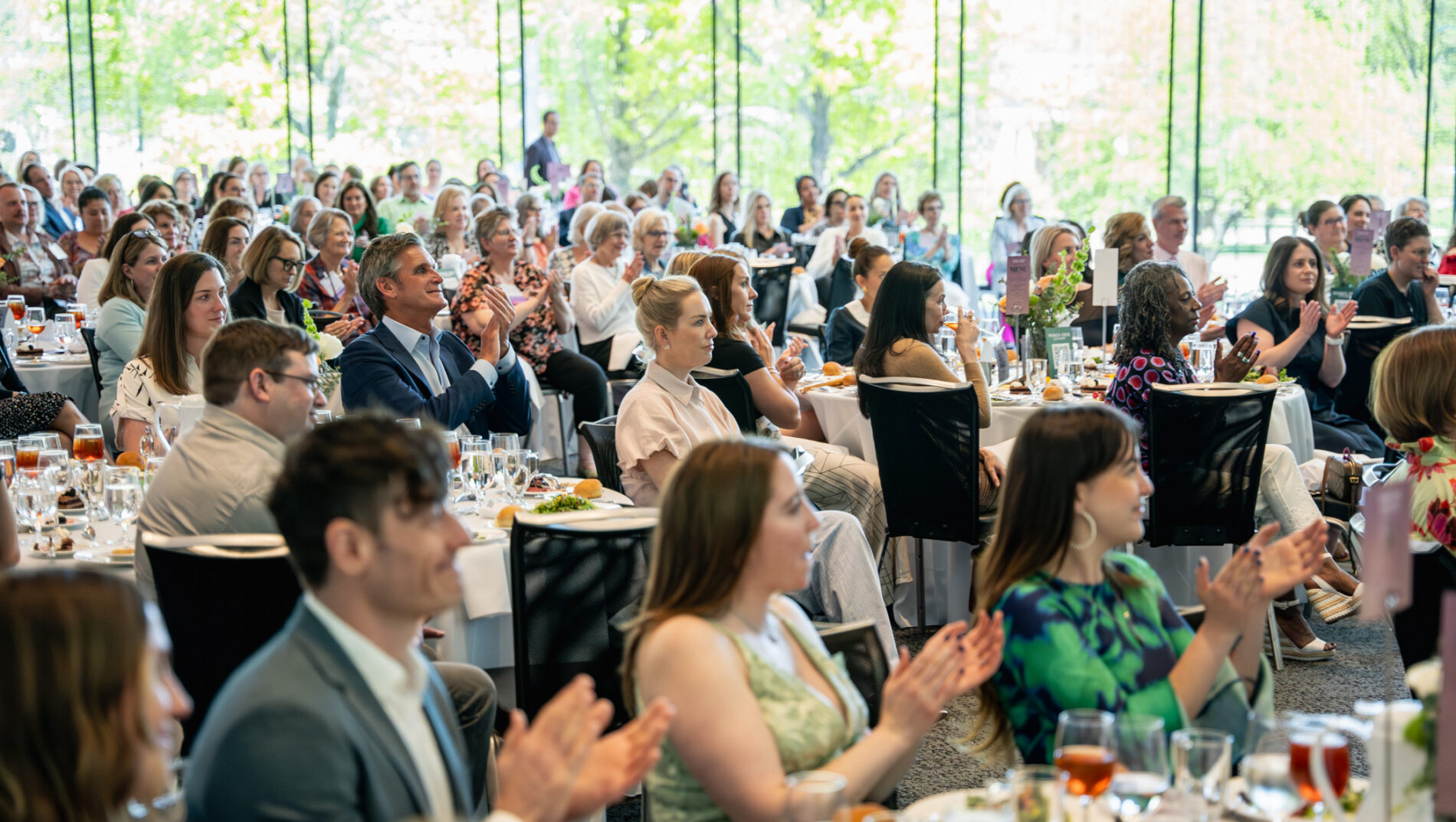 Audience clapping and engaged during a formal dining event with large windows showing greenery outside