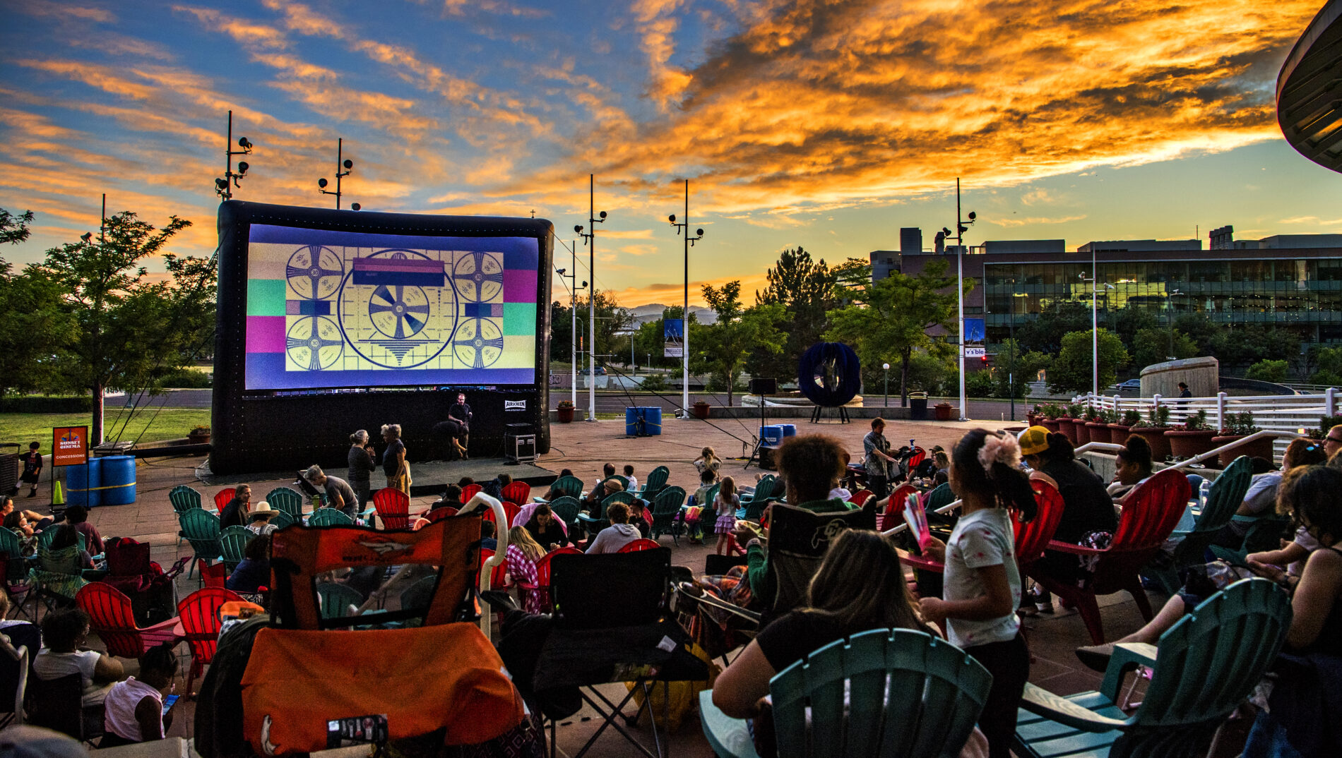 Outdoor movie screen showing test pattern, colorful sunset, and audience seated in chairs