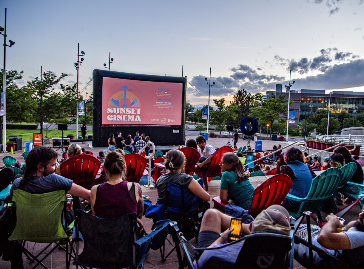 Outdoor movie screening at sunset with audience in camping chairs. Large screen displays "Sunset Cinema" and event details
