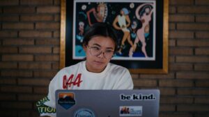 KAMNOP-3.jpg person focusing on laptop with stickers, colorful artwork behind