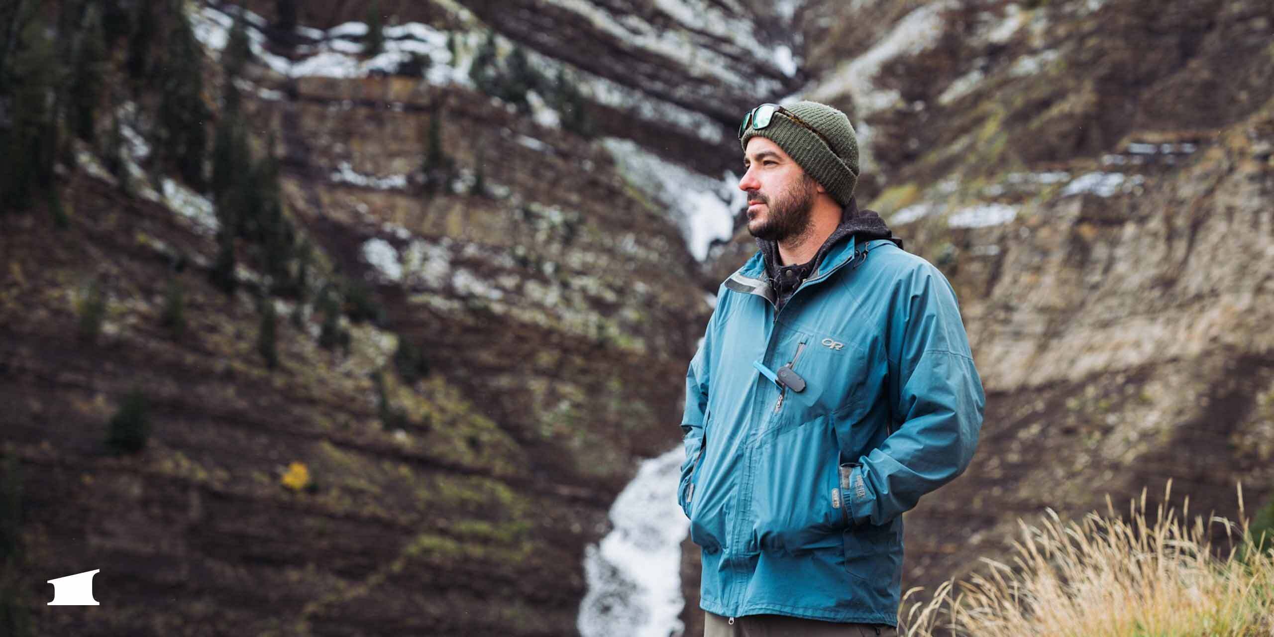 Man in blue jacket and green beanie standing near waterfall and snowy cliff