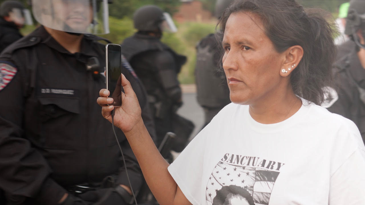 Woman stands holding a phone, facing police officers in riot gear, wearing a shirt with the word "Sanctuary."