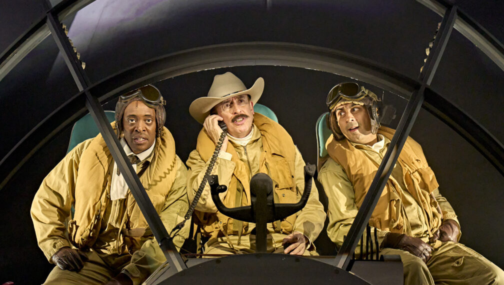 Three people in a cockpit, wearing yellow flight suits and headgear. Central figure wears a cowboy hat, holding a radio