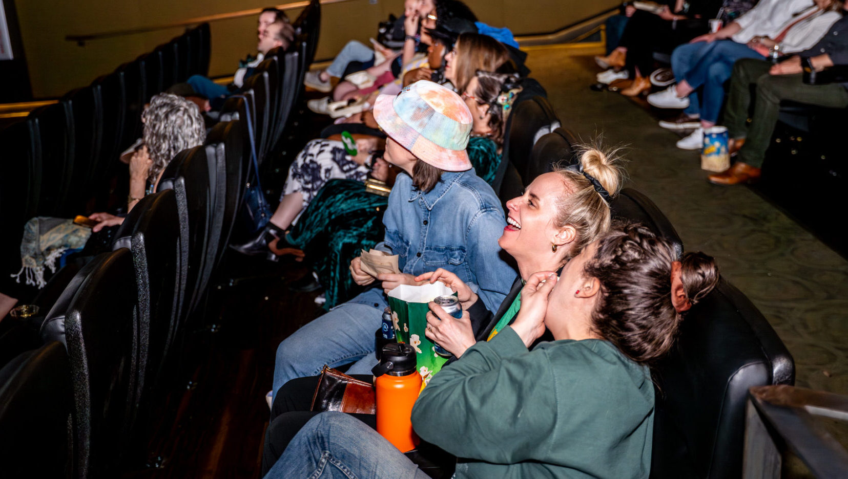 Audience in a theater laughing, holding popcorn and drinks, enjoying a screening